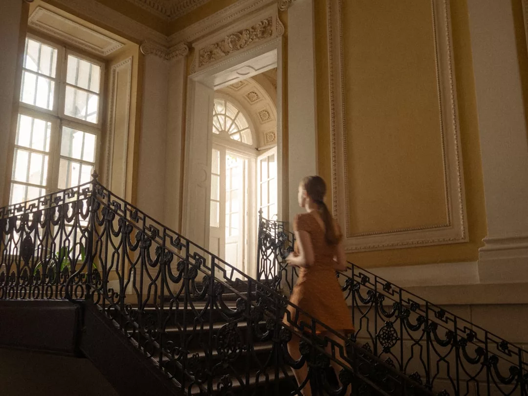 Una mujer con un vestido marrón sube por una escalera decorativa en un edificio con grandes ventanales y detalles arquitectónicos ornamentados, que evocan el lujo intemporal de Mallorca.