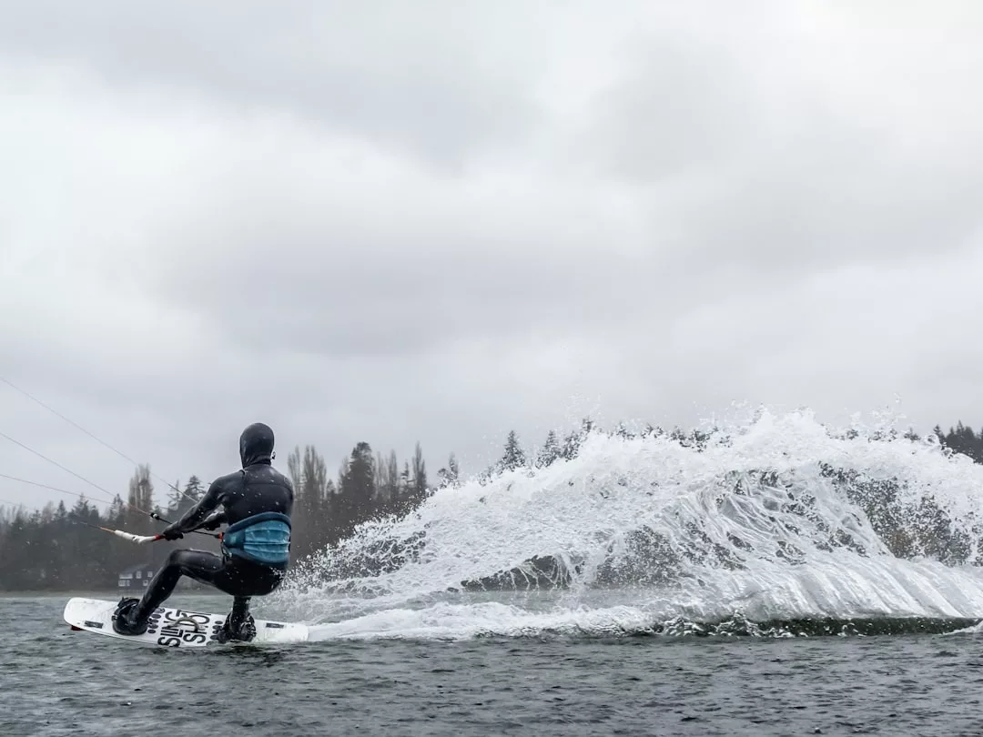 Una persona con traje de neopreno practica wakeboard en un lago de Mallorca, creando un gran chapoteo a sus espaldas bajo un cielo nublado.