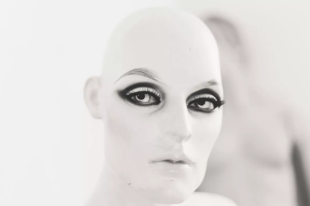 Un primer plano de la cabeza de un maniquí calvo con un espectacular maquillaje negro en los ojos capta la esencia de Lujo, con otro maniquí difuminado al fondo, que evoca el escaparate de una elegante boutique de Mallorca.
