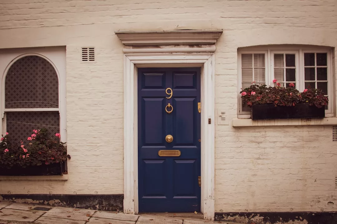Una puerta azul con el número 9 y una ranura de latón para el correo está encajada en una pared de ladrillo blanco, flanqueada por dos ventanas con jardineras negras llenas de flores rosas, que evocan el lujo y el encanto de las casas con estilo de Mallorca.