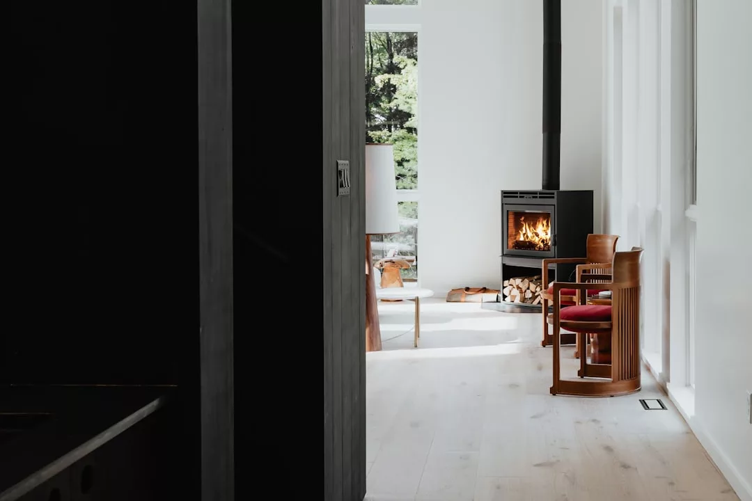 Un moderno salón con suelos de madera clara, dos sillas de madera y una estufa de leña desprende lujo, mientras que la leña apilada y los grandes ventanales revelan la belleza natural de Mallorca en el exterior.