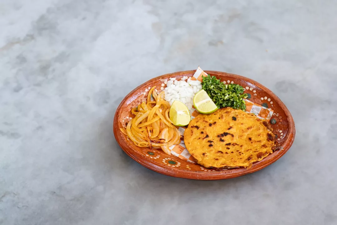 Un plato con una tortilla cocida, cebollas asadas, gajos de lima, cebollas picadas y cilantro picado sobre una superficie gris aporta un toque de Lujo a su mesa, perfecto para saborear sabores inspirados en Mallorca.