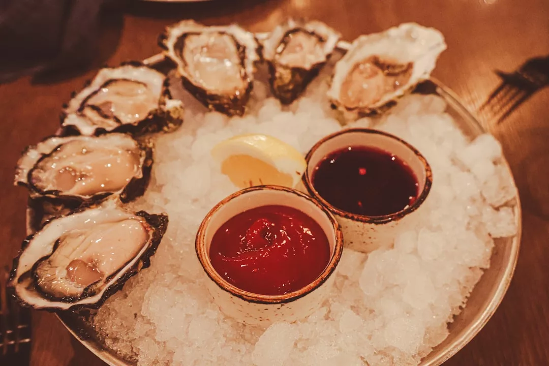 Un plato de seis ostras crudas en su media concha, servidas sobre hielo picado con una rodaja de limón y dos pequeños cuencos de salsa de cóctel y mignonette, un verdadero sabor de lujo que recuerda la elegancia costera de Mallorca.