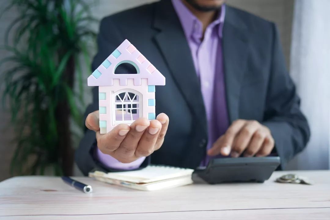 Una persona trajeada sostiene una pequeña maqueta de casa en una mano y utiliza una calculadora con la otra, con un bolígrafo, un cuaderno y unas llaves sobre la mesa, captando a la perfección la esencia del lujo inmobiliario en Mallorca.