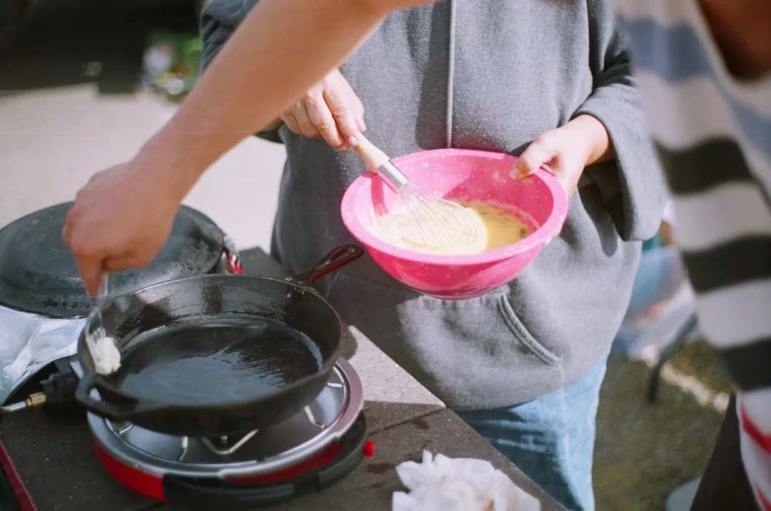 Dos personas disfrutan de un toque de lujo en Mallorca mientras preparan comida al aire libre; una bate masa en un cuenco rosa mientras la otra unta aceite en una sartén de hierro fundido sobre un hornillo portátil.