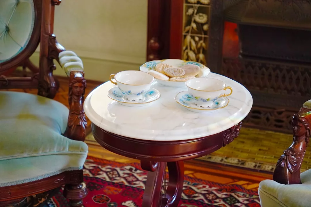 Una mesa redonda de mármol con dos tazas de té, platillos y un plato de galletas descansa entre dos sillas tapizadas ornamentadas, evocando el relajado lujo de Mallorca.
