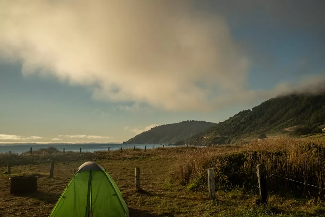 Se instala una carpa verde en un terreno cubierto de hierba cerca de la costa de Mallorca, con las colinas y el océano de fondo bajo un cielo parcialmente nublado.