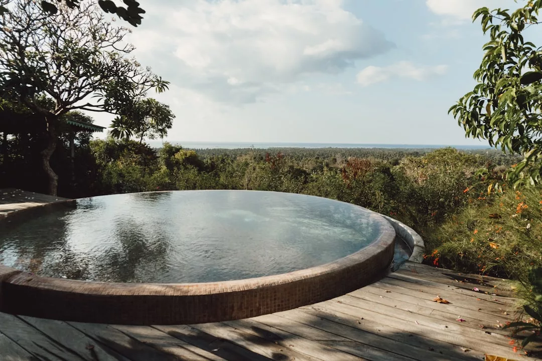 Piscina infinita circular de aguas tranquilas situada en una terraza de madera en Lujo Mallorca, con vistas a frondosos árboles verdes y un horizonte lejano bajo un cielo parcialmente nublado.