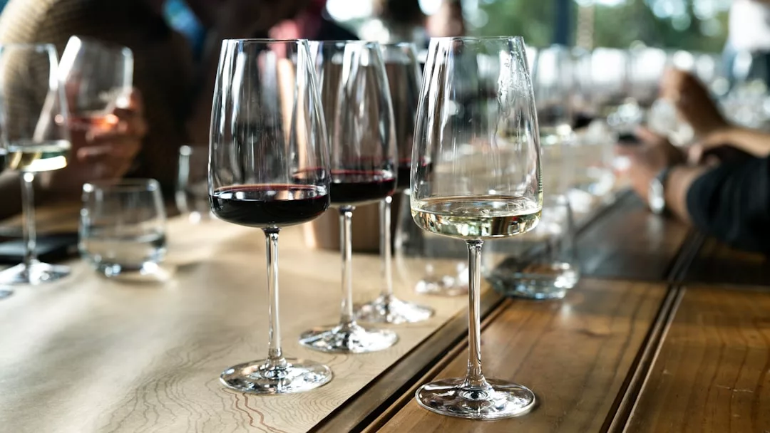 Una hilera de copas con vino tinto y blanco sobre una mesa de madera, rodeada de gente al fondo, desprende un aire de lujo que recuerda a las sofisticadas reuniones de Mallorca.
