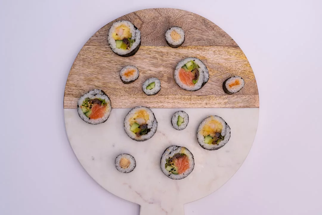 Una tabla de cortar redonda con secciones de madera y mármol muestra rollos de sushi variados con diversos rellenos, dispuestos de forma dispersa, aportando un toque de lujo mallorquín a su experiencia gastronómica.