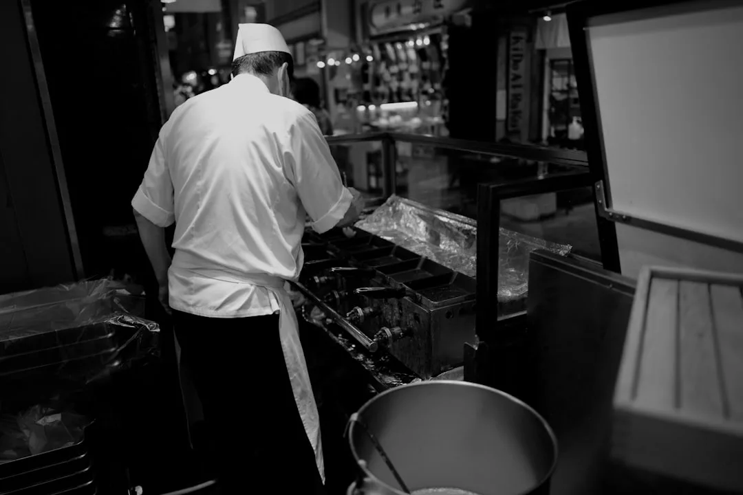 Un chef con uniforme y delantal blancos cocina a la parrilla en una cocina comercial, visto desde atrás en una foto en blanco y negro que capta el tranquilo lujo de la escena culinaria de Mallorca.