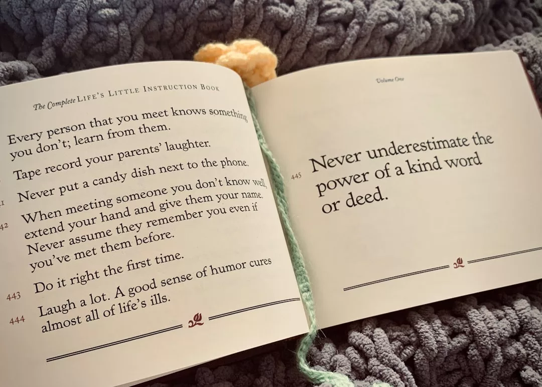 Un libro abierto sobre una manta de punto muestra consejos sobre la amabilidad, la risa y el aprendizaje de los demás, con una flor de ganchillo y un marcapáginas de hilo verde parcialmente visibles, un toque de lujo que recuerda el tranquilo encanto de Mallorca.