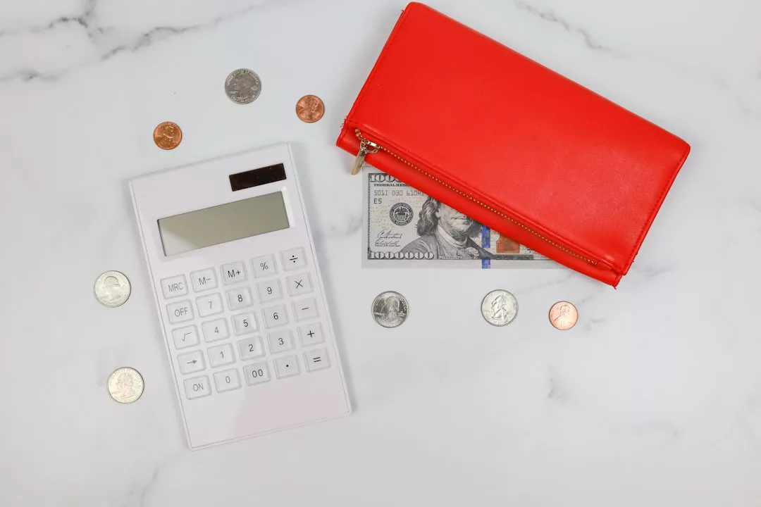 Una calculadora blanca, monedas variadas, un billete de 100 dólares y un monedero rojo con cremallera evocan un toque de lujo mientras están colocados sobre una superficie blanca, como si estuvieras contando los gastos antes de una escapada a Mallorca.