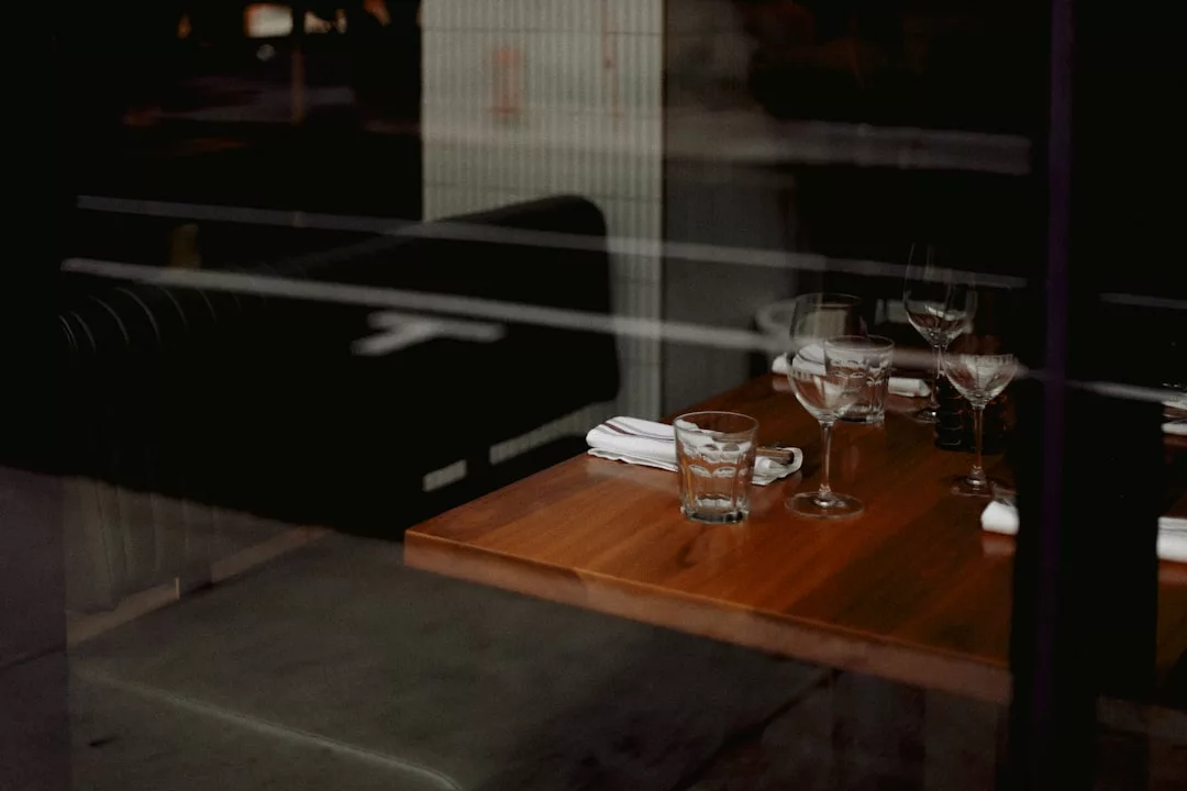 Una mesa de restaurante de madera está puesta con vasos, servilletas y cubiertos, vista a través de una ventana desde el exterior, capturando el relajado lujo de cenar en Mallorca.