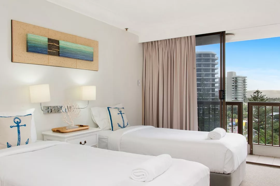 Una luminosa habitación de hotel en Mallorca con dos camas individuales, almohadas de temática náutica, una mesa auxiliar con decoración de coral y un balcón con vistas a la ciudad y al mar, perfecta para una relajante escapada de lujo.