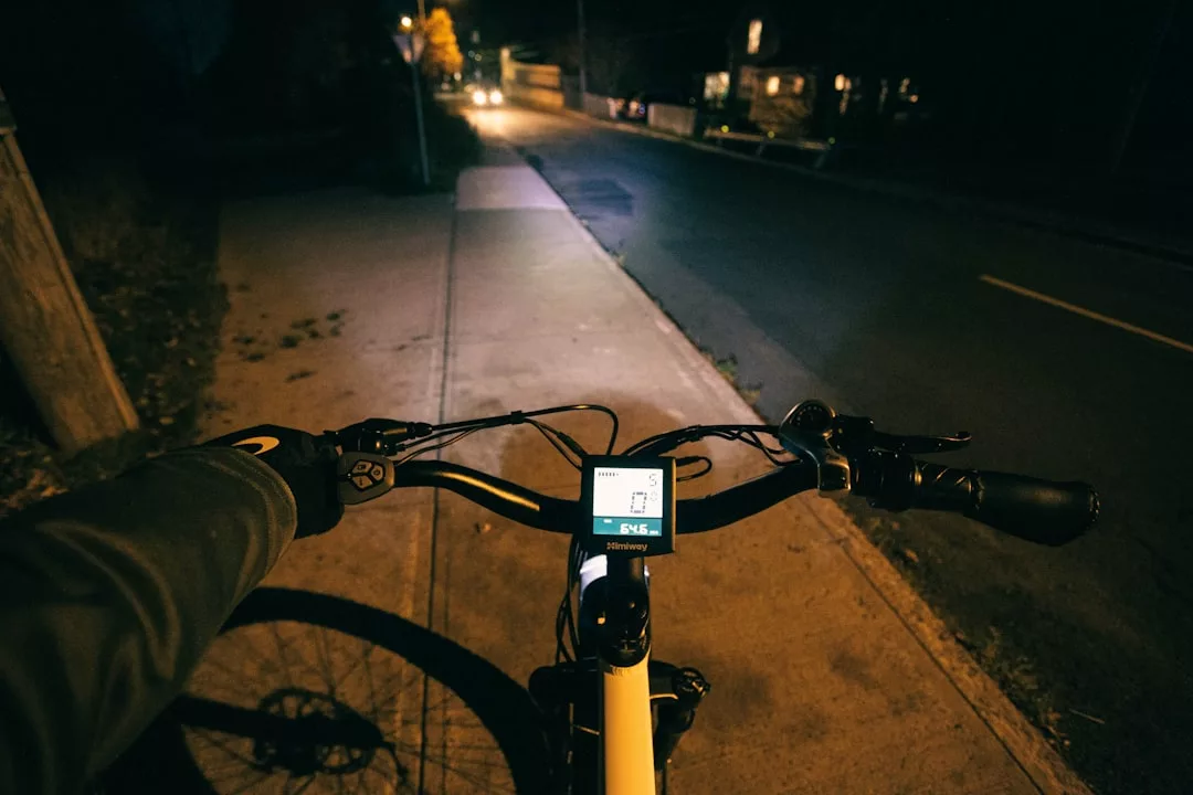 Vista desde detrás del manillar de una bicicleta por la noche en Mallorca, mostrando una pantalla de visualización encendida y una acera poco iluminada con farolas y un coche que se acerca, captando el tranquilo lujo de montar en bicicleta al anochecer.