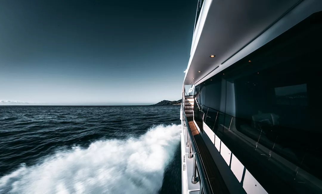 Vista desde la cubierta lateral de un yate en movimiento en aguas abiertas cerca de Mallorca, con olas blancas que se arrastran detrás y una costa distante visible bajo un cielo despejado, capturando la esencia del lujo en el mar.