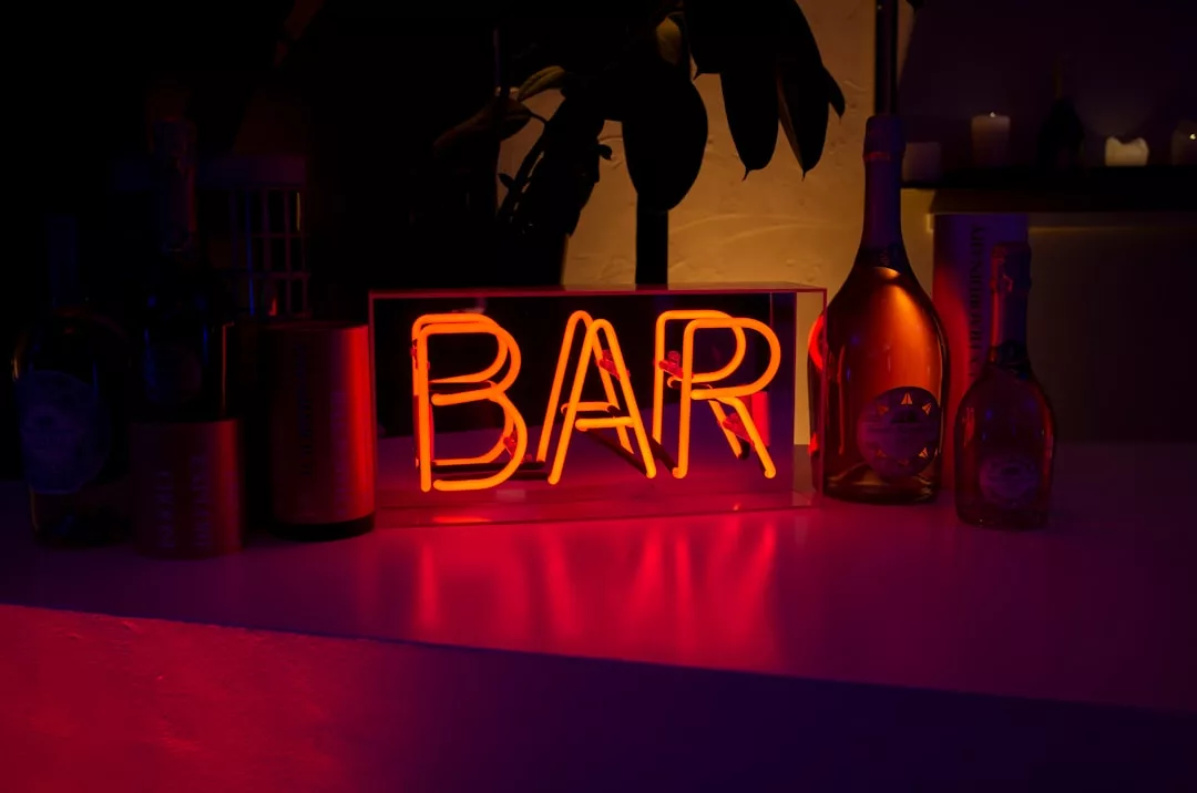 Un brillante letrero de neón que reza "BAR" está rodeado de botellas sobre una superficie poco iluminada, evocando el lujo de las noches mallorquinas, con sombras oscuras y la tenue luz de las velas de fondo.