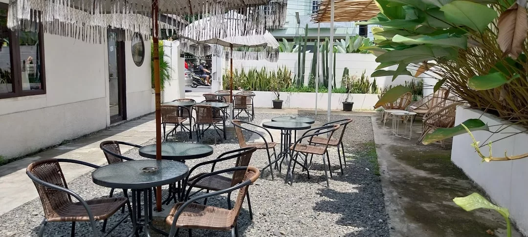 Zona de asientos al aire libre con mesas redondas de cristal y sillas de mimbre sobre grava, a la sombra de sombrillas, rodeada de frondosas plantas y una pared blanca: un oasis de lujo que recuerda la relajada elegancia de Mallorca.