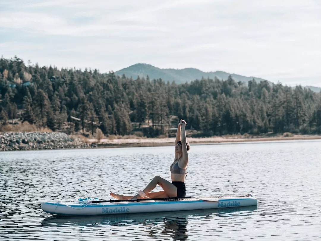 Una persona vestida con ropa deportiva se sienta en una tabla de paddle surf en un lago de Mallorca, estirando los brazos por encima de la cabeza, con colinas boscosas y árboles que aumentan la sensación de lujo y tranquilidad.