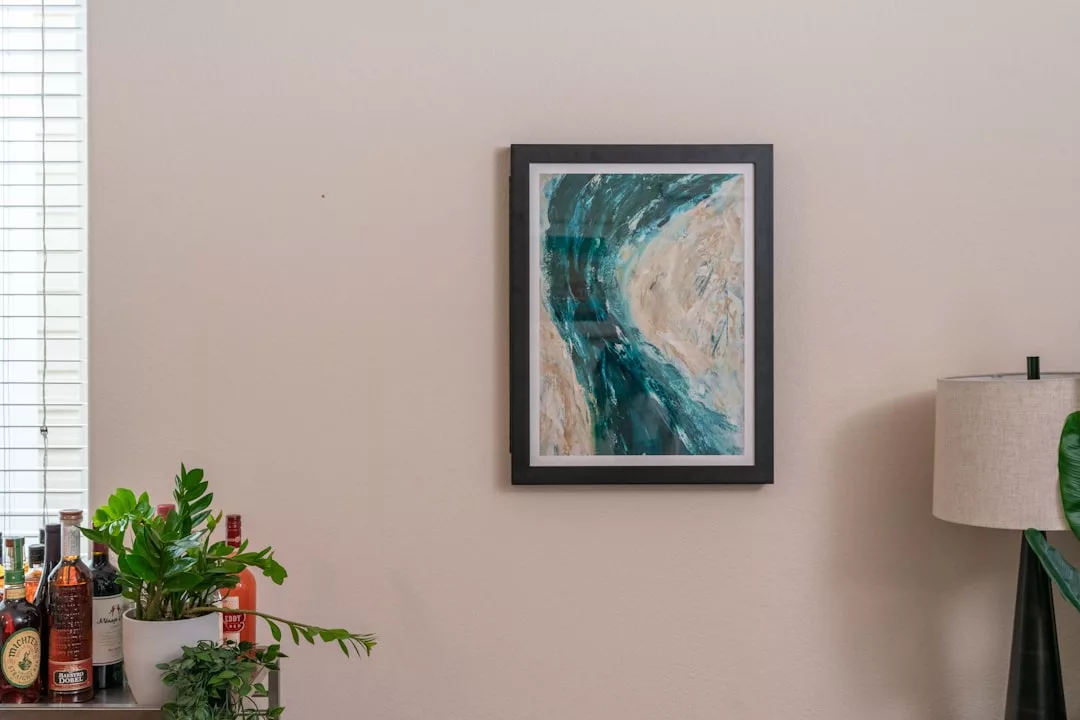 Un cuadro abstracto enmarcado en tonos azules y beige cuelga de una pared beige, añadiendo un toque de lujo sobre un carrito de bar con plantas, botellas y una lámpara cerca, evocando la relajada elegancia de Mallorca.