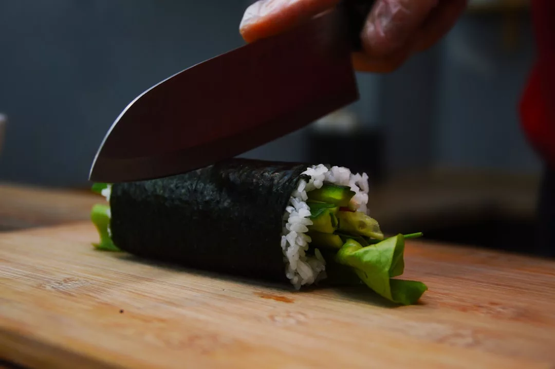 Una mano sostiene un cuchillo preparado para cortar un rollo de sushi relleno de arroz y verduras sobre una tabla de cortar de madera, capturando un momento de una cena inspirada en el lujo en Mallorca.